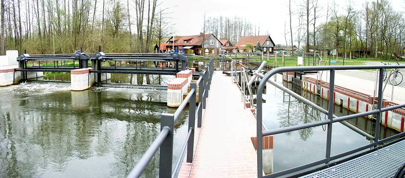 Die Wehrh&ouml;he ist einstellbar, damit kann der Wasserstand der vorgelagerten Flie&szlig;e reguliert werden. Im Hintergrund sind die Geb&auml;ude des Leiper Campingplatzes zu sehen.